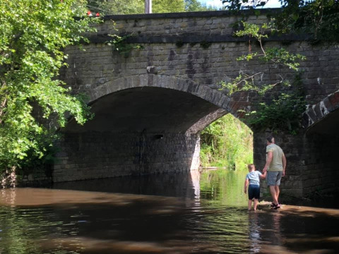 A man and child walk through shallow water under an old stone bridge near Nassogne, Belgium.