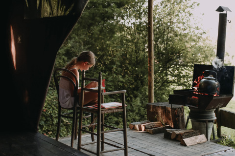 A girl sits at a table on a wooden deck at Feather Down Rochefort-Ardenne, reading a book near a kettle.