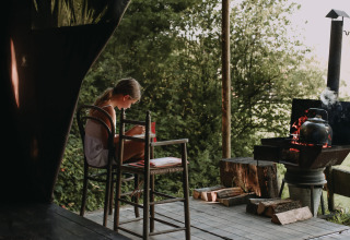 Een meisje leest een boek op een houten terras bij Feather Down Rochefort-Ardenne, naast een ketel.