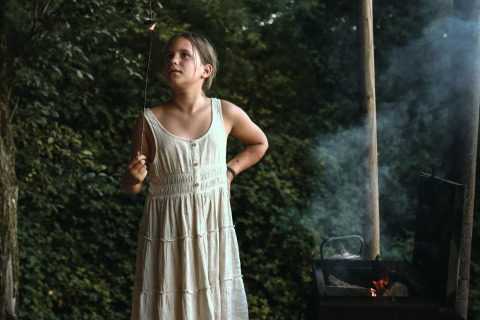 Una ragazza in abito bianco tosta marshmallow su un fuoco da campo a Feather Down Rochefort-Ardenne.