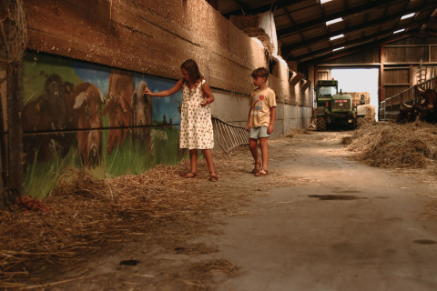 Dos niños juegan en un granero con heno en Feather Down Rochefort-Ardenne, Bélgica, con un tractor al fondo.