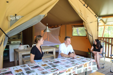 Tre persone si rilassano all'interno di una tenda safari con arredi in legno e luce naturale esterna.