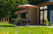 Terrasse der Cannenburch-Lodge im Veluwepark de Bosgraaf, Niederlande, mit Tisch, Stühlen und Sonnenschirm.