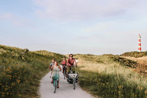 Familia pedaleando por un sendero arenoso cerca de Hollum, Frisia, con un faro rojo y blanco al fondo.