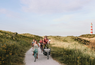 Familia pedaleando por un sendero arenoso cerca de Hollum, Frisia, con un faro rojo y blanco al fondo.