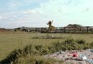 Bambina con vestito giallo che salta sull’erba al Feather Down Boerderij Ameland, Friesland, Paesi Bassi.
