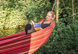 Mujer relajándose en una hamaca junto a un pod, leyendo un libro, rodeada de vegetación y árboles.