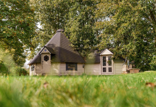 Alojamiento glamping acogedor con cabaña de madera, rodeado de césped verde y grandes árboles frondosos.
