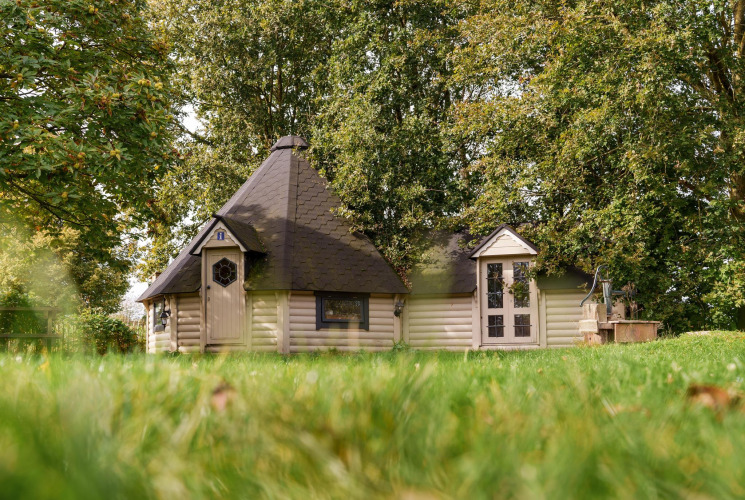 Alojamiento glamping acogedor con cabaña de madera, rodeado de césped verde y grandes árboles frondosos.