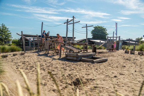 Children and adults play at a sandy playground near the Berkel cabin on a sunny day, surrounded by cabins.