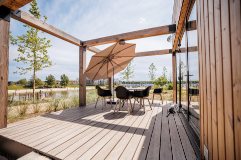 Terraza de madera de una cabaña moderna con mesa, sillas y sombrilla, vista al lago y la playa.
