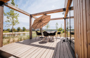 Houten lodge-terras met moderne stoelen en parasol, uitzicht op strand en water in de verte.