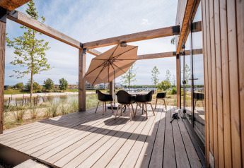 Terraza de madera de una cabaña moderna con mesa, sillas y sombrilla, vista al lago y la playa.