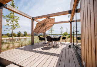 Houten lodge-terras met moderne stoelen en parasol, uitzicht op strand en water in de verte.