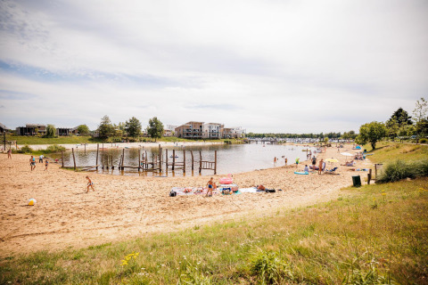 Foto de una playa de arena en un lodge con personas relajándose y jugando junto al lago en un día soleado.