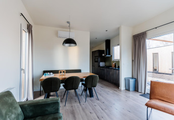 Eetruimte in een moderne lodge met groene stoelen en grote ramen, met een open keuken op de achtergrond.