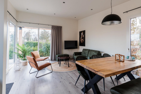 Salon moderne de lodge avec grandes fenêtres, canapé vert, fauteuil en cuir, table en bois et vue jardin.