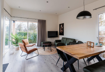 Moderne lodge-living met grote ramen, groene zetel, lederen stoel, houten tafel en zicht op tuin.