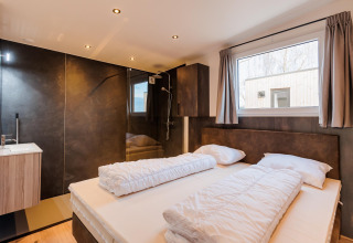 Dormitorio moderno con cama doble, ventana grande y baño privado en Beachlodge Deluxe en Vakantiepark Leukermeer.