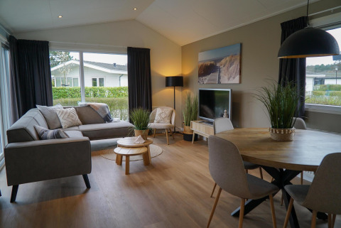 Modernes Wohnzimmer in der Velthorst-Hütte mit grauem Sofa, Esstisch, Pflanzen und großen Fenstern.