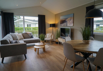 Sala moderna en la cabaña Velthorst con sofá gris, mesa redonda, plantas y grandes ventanales con vista.