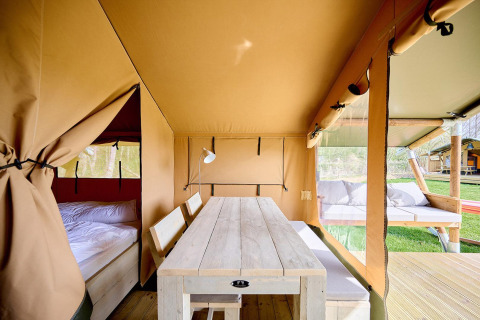 Interior de una tienda safari Villatent Compact con mesa de madera, banco, cama y zona lounge exterior.