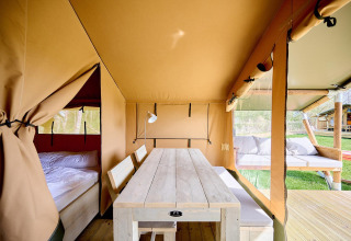 Interior de una tienda safari Villatent Compact con mesa de madera, banco, cama y zona lounge exterior.