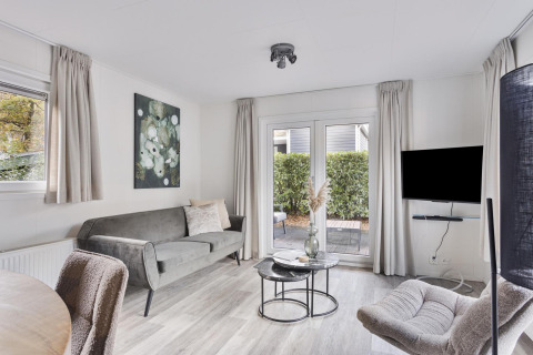Sala de estar moderna y luminosa con sofá gris, sillones, televisor y acceso al patio en Wellness Lodge, Países Bajos.