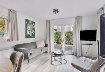 Sala de estar moderna y luminosa con sofá gris, sillones, televisor y acceso al patio en Wellness Lodge, Países Bajos.