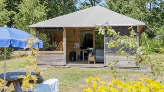 Cabaña de madera Camphome con porche cubierto y hamaca, rodeada de árboles y situada sobre césped.