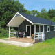 Chalet med veranda omgivet af grønt græs og træer, mennesker nyder udendørs siddepladser i solen.