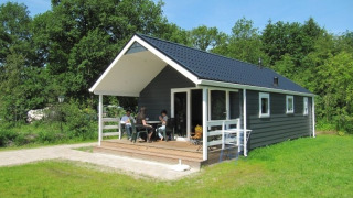 Chalet med veranda omgivet af grønt græs og træer, mennesker nyder udendørs siddepladser i solen.