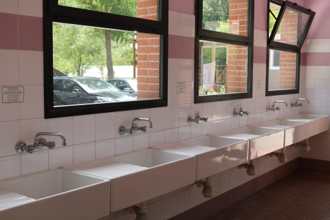 Hilera de lavabos blancos bajo ventanas abiertas en el baño común de Camping Il Poggetto, Italia.