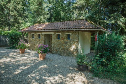 Stone building with a tiled roof at Camping Lestaubière in France, surrounded by trees and potted flowers.