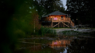 Safari tent cottage on stilts lit up at dusk by a pond at Holiday Park Sallandshoeve, Netherlands.