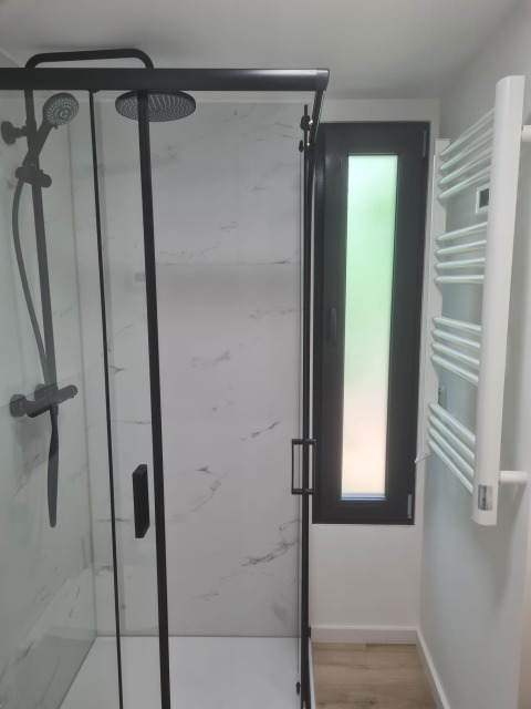 Ducha moderna con puerta de cristal, rociador tipo lluvia, marco negro y ventana estrecha en tiny house.