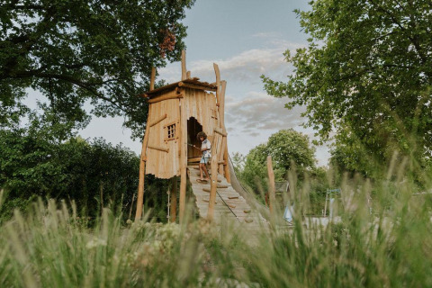 Un niño juega en una cabaña de madera en un sitio de glamping rodeado de vegetación y árboles altos.
