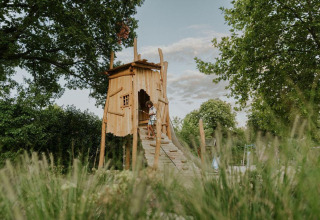 Et barn leger i et charmerende træhus omgivet af natur på en glampingplads under en blå himmel.