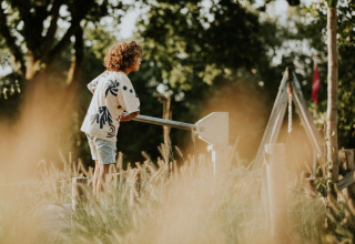 Een kind staat aan een leuning tussen hoog gras bij een glampingverblijf midden in de natuur.