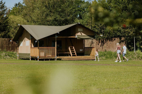Familie går forbi Ranger-lodge på Camping Si-Es-An i Holland, omgivet af natur og frodigt grønt.