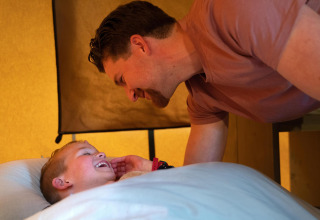 Een vader leunt over zijn lachende zoon in bed in Ranger Lodge op Camping Si-Es-An in Nederland.