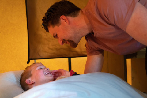 Ein Vater beugt sich über seinen lachenden Sohn im Bett in der Ranger Lodge auf Camping Si-Es-An, Niederlande.