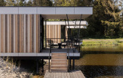 Moderne glampingaccommodatie boven water met veranda, houten gevelbekleding en uitzicht op natuur.
