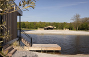 Cabaña de madera junto a un lago con embarcadero, playa y tienda glamping rodeada de bosque.