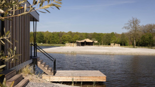 Wooden cabin with steps leading to a dock by a lake, sandy beach and glamping tent in a forested area.