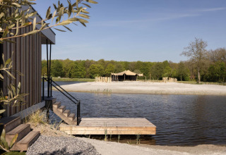 Træhytte ved sø med udsigt til strand og telt, omgivet af skov, perfekt til glampingoplevelser.