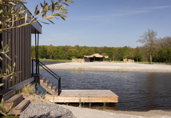 Træhytte ved sø med udsigt til strand og telt, omgivet af skov, perfekt til glampingoplevelser.