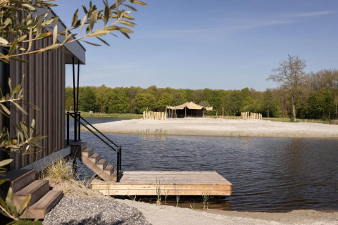 Cabina in legno sul lago con pontile, spiaggia sabbiosa e tenda glamping immersi nel verde bosco.