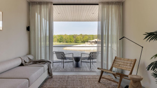 Minimalist glamping accommodation living room with sofa, wood chair and lake view through glass doors.