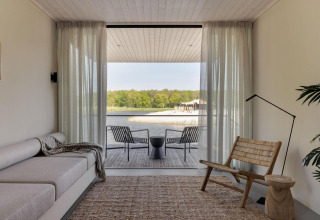 Sala de estancia minimalista en glamping con terraza y vistas al lago y bosque a través de puertas de cristal.